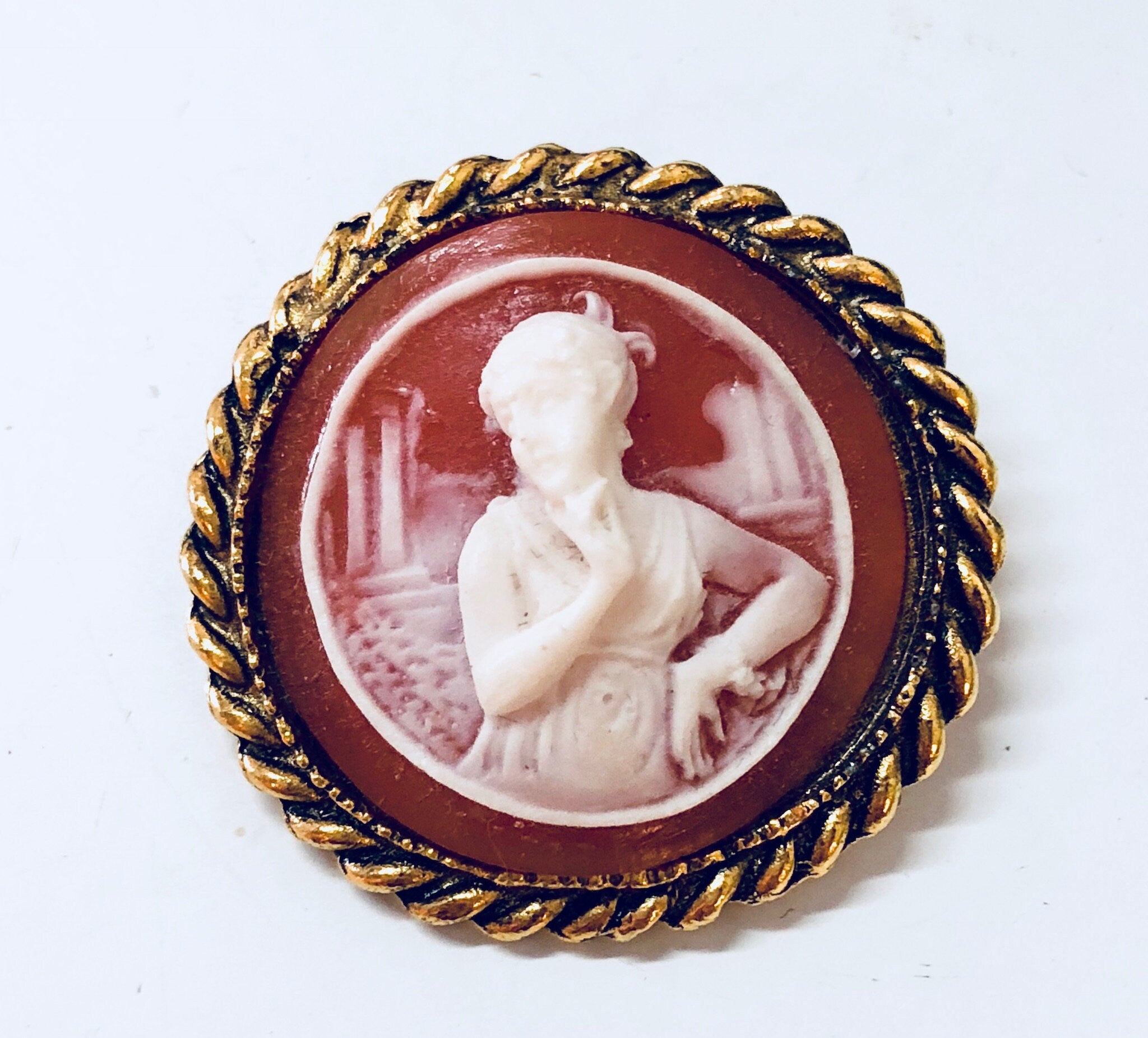 Vintage gold-toned cameo brooch featuring a white carved figure against a dark orange background, surrounded by a twisted rope border.