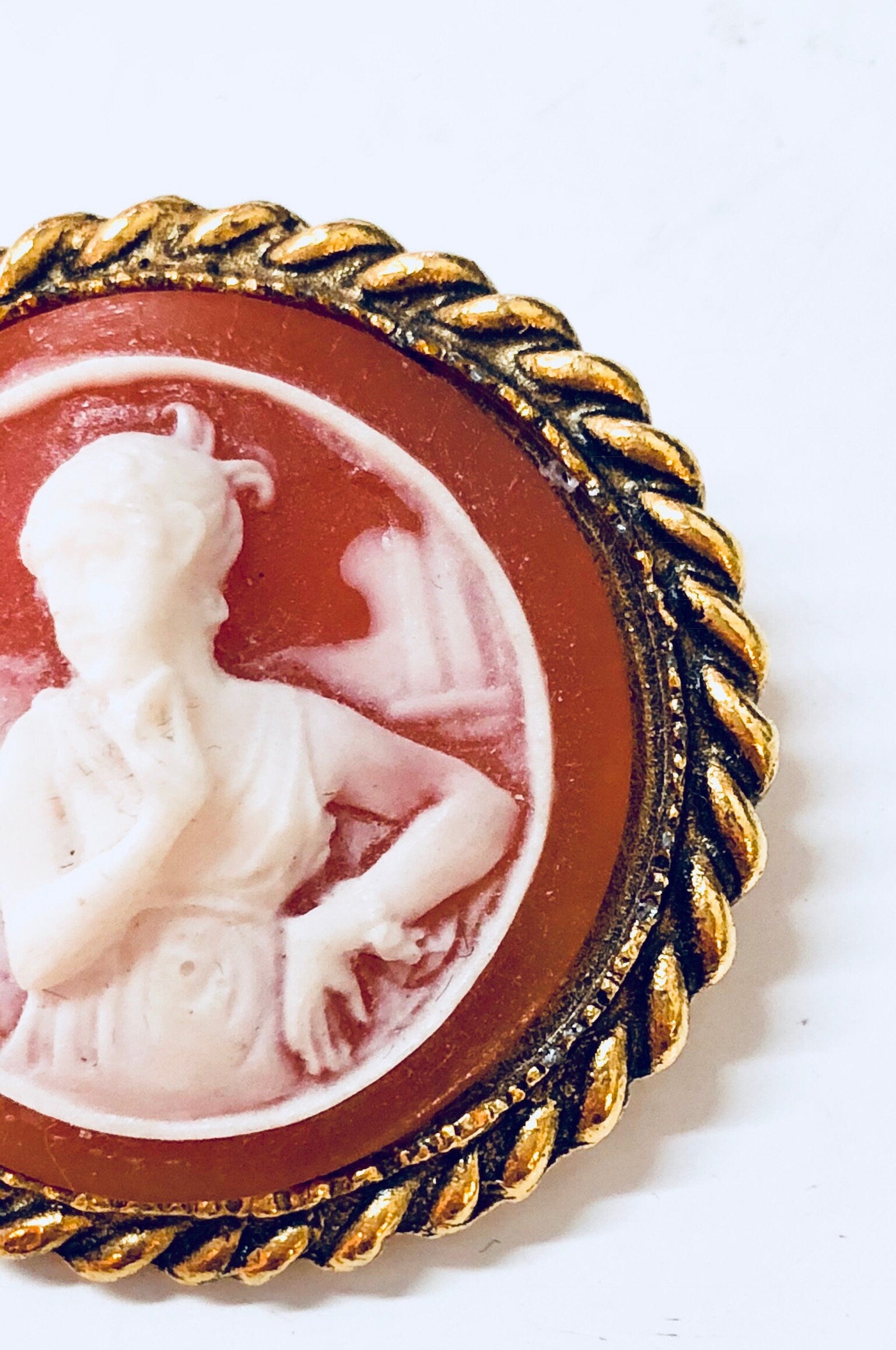 Vintage gold-toned cameo brooch featuring a white carved figure on an orange background, surrounded by a detailed rope-style border.
