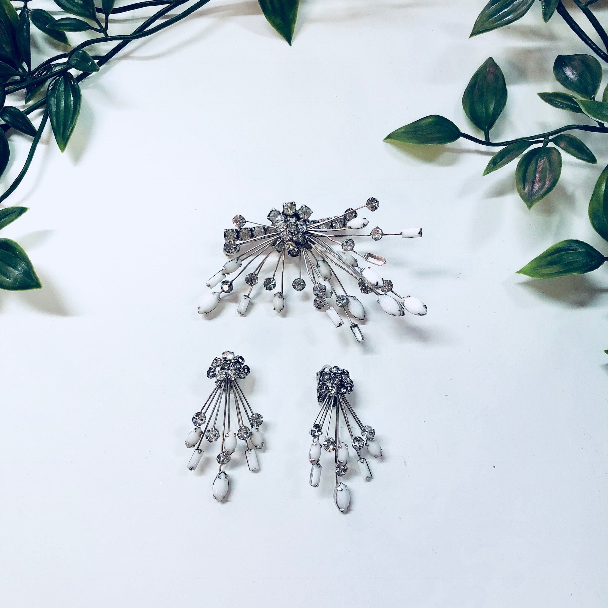 Silver Art Deco style Marvella jewelry set featuring a brooch and clip-on earrings with white rhinestones arranged in a starburst design, displayed on a white background with green leaves.