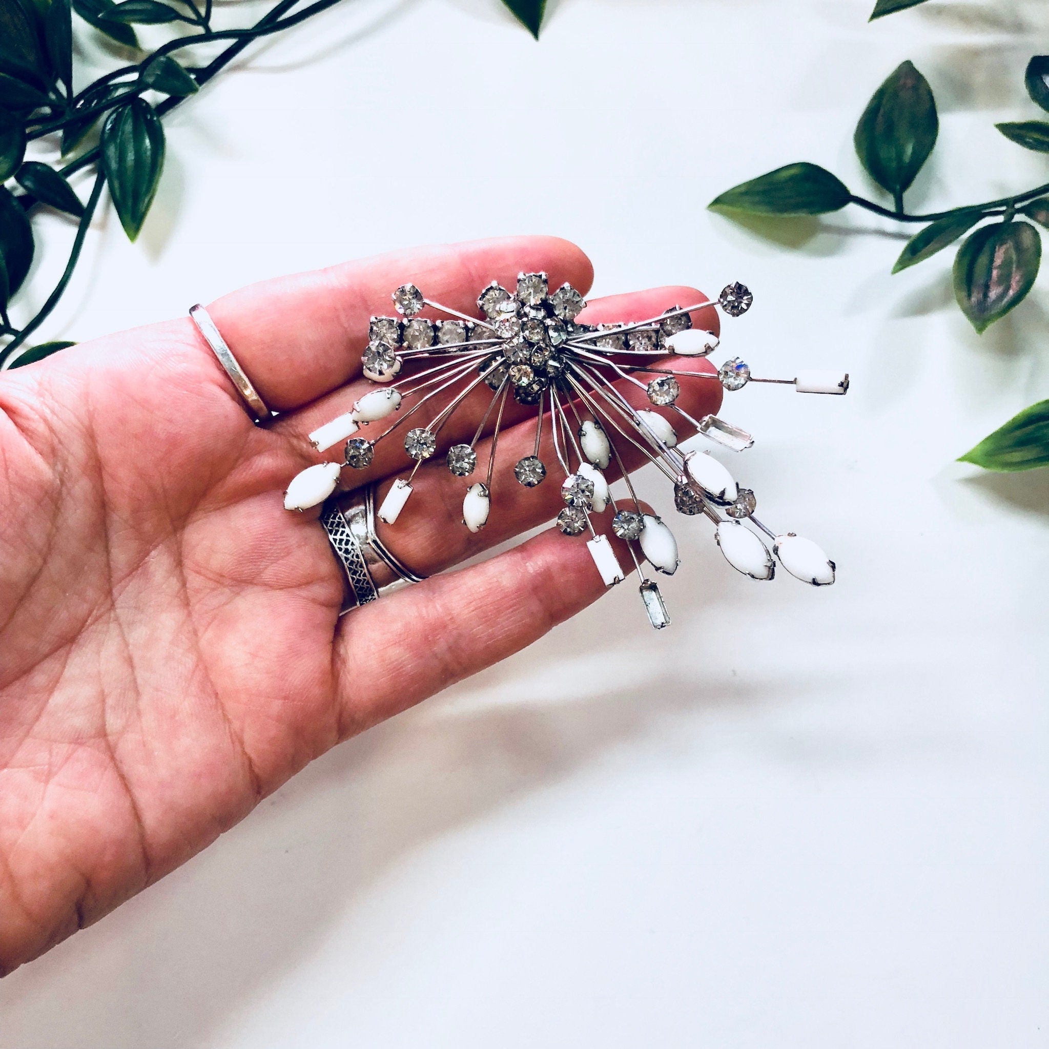 Marvella jewelry set with a sparkling rhinestone brooch and matching clip-on earrings featuring unique white stones and an art deco silver design, held in a hand against a white fabric background with greenery.
