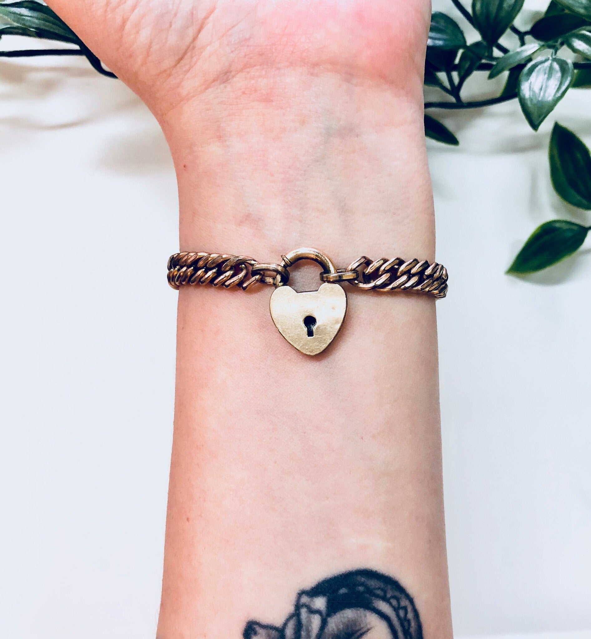 Vintage gold-toned chain bracelet with heart-shaped locket charm on a tattooed wrist, with plant in background.