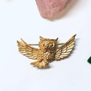 Vintage gold-toned owl brooch with spread wings and textured details, displayed on a white background near a rose quartz crystal.