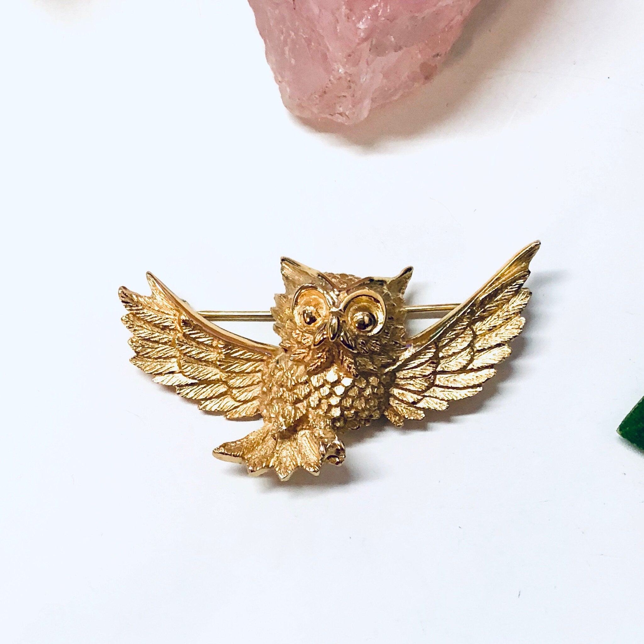 Vintage gold-toned owl brooch with spread wings and textured details, displayed on a white background near a rose quartz crystal.