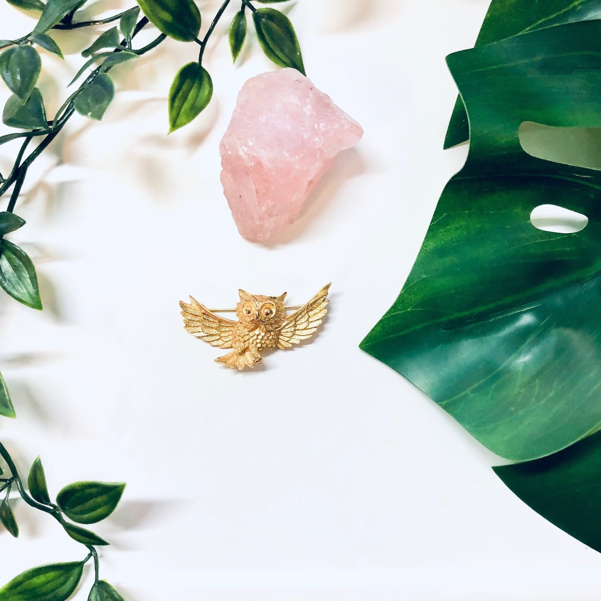 Vintage gold-toned owl brooch by Trifari displayed on white fabric with pink quartz crystal and green leaves.