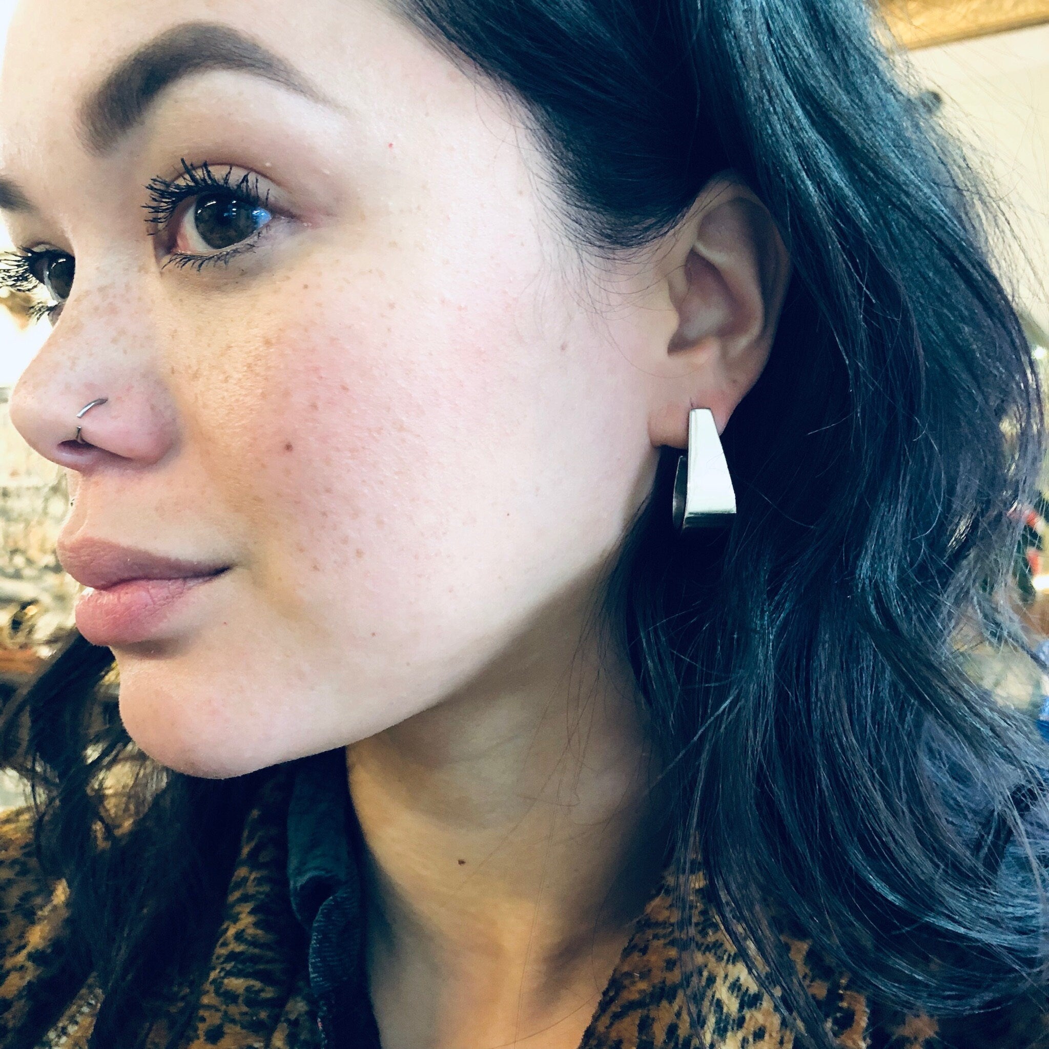 The image shows a close-up of a woman's face. She has fair skin with some freckles, green eyes, and dark wavy hair. The woman is wearing triangular white earrings and an animal print top or scarf. She appears to be looking off to the side with a slight smile.