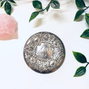 Vintage sterling silver compact mirror with intricate floral motif and monogram, surrounded by green leaves, on a white fabric background.