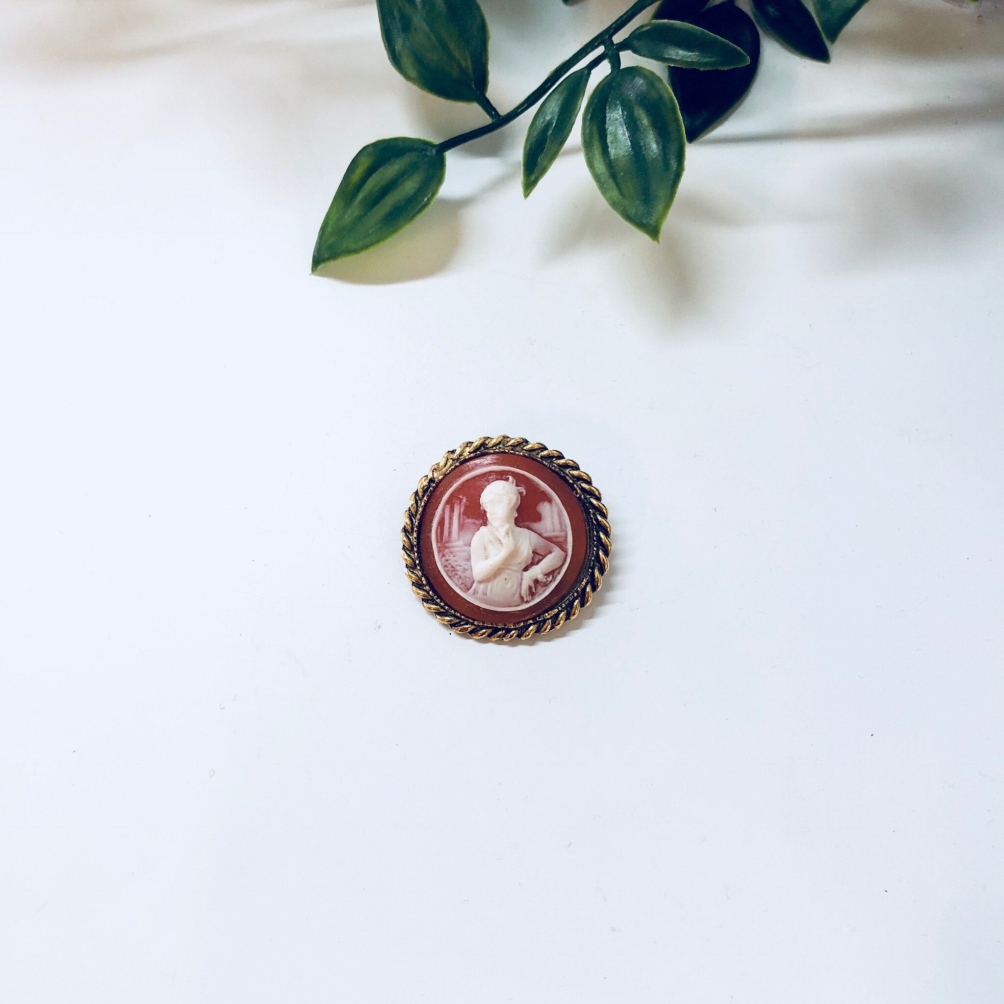 Vintage gold-toned cameo brooch featuring an orange and white Victorian-style portrait, displayed on a white fabric surface with green foliage in the background.