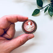 Vintage cameo brooch with a white carved profile set in an ornate gold-toned frame, held in a person's hand against a white background with green foliage.