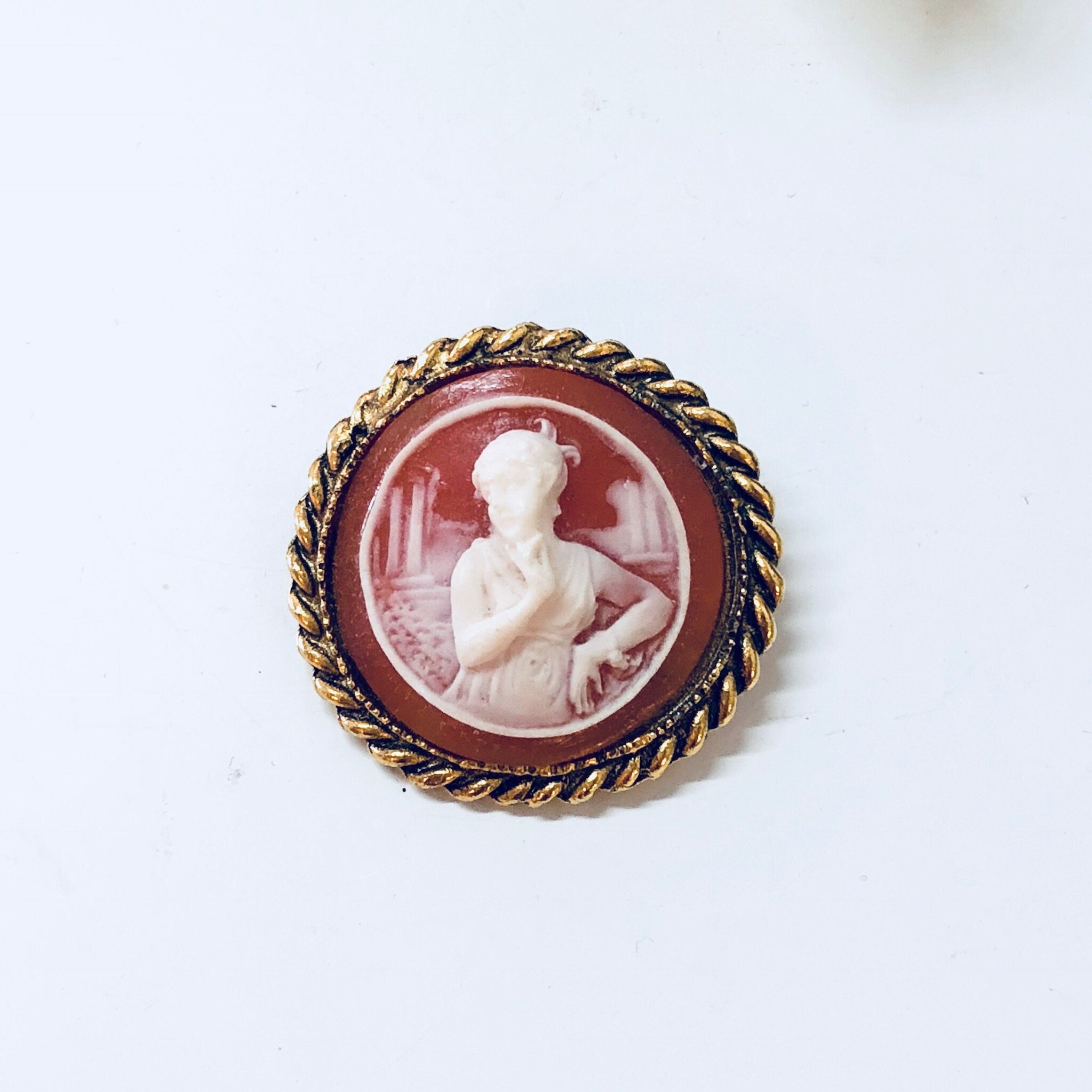 Vintage gold-toned cameo brooch featuring a carved shell portrait of a woman in profile set against an orange background, encircled by a twisted rope border.