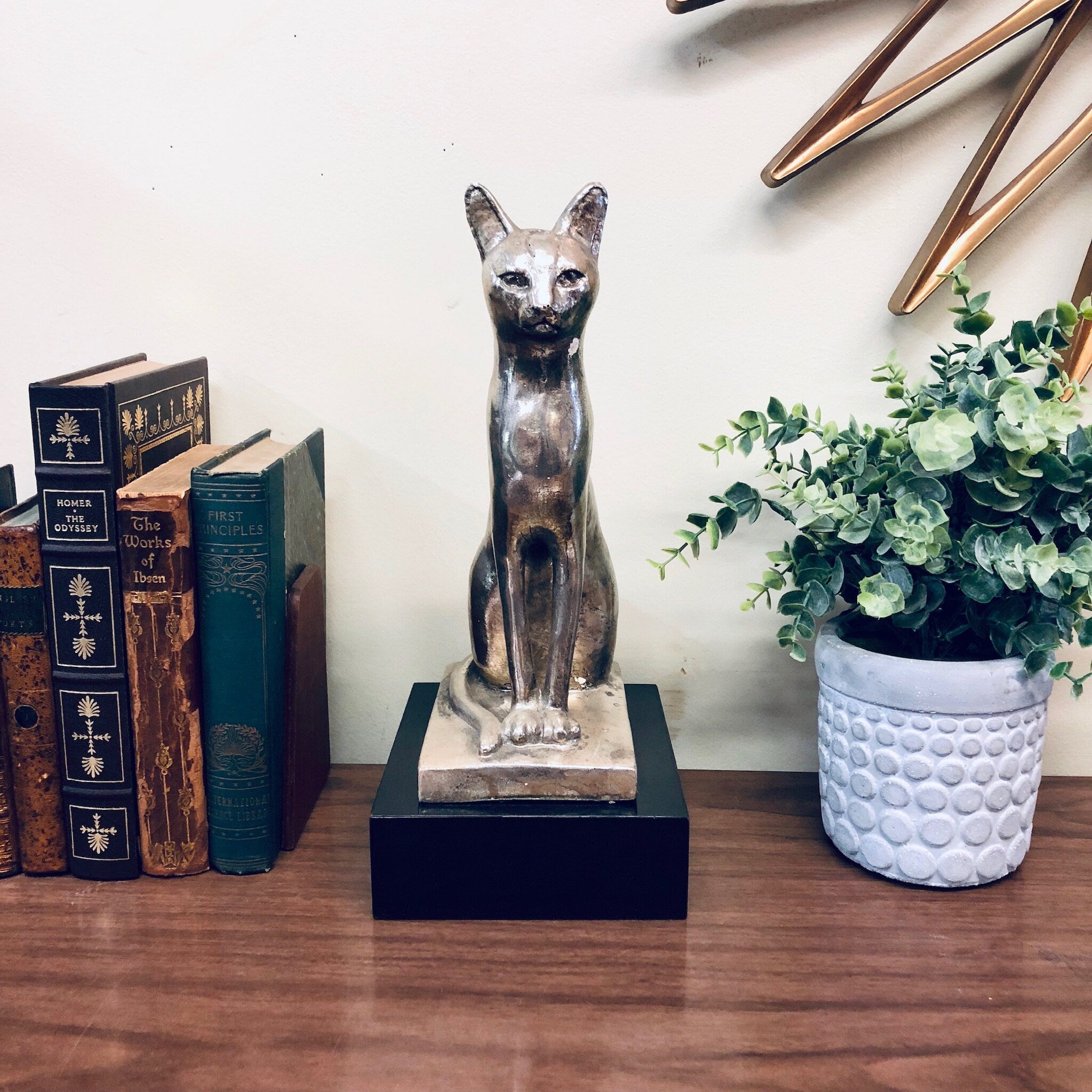 Vintage silver cat sculpture displayed on wooden surface with books and potted plant, serving as unique home decor piece.