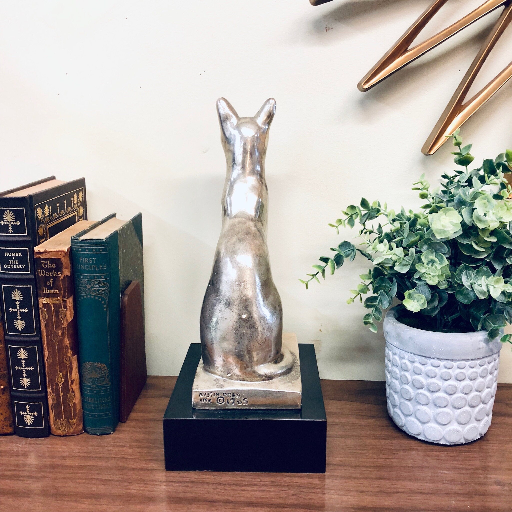 Silver cat sculpture displayed on black base surrounded by books and plant, vintage home decor on wooden surface