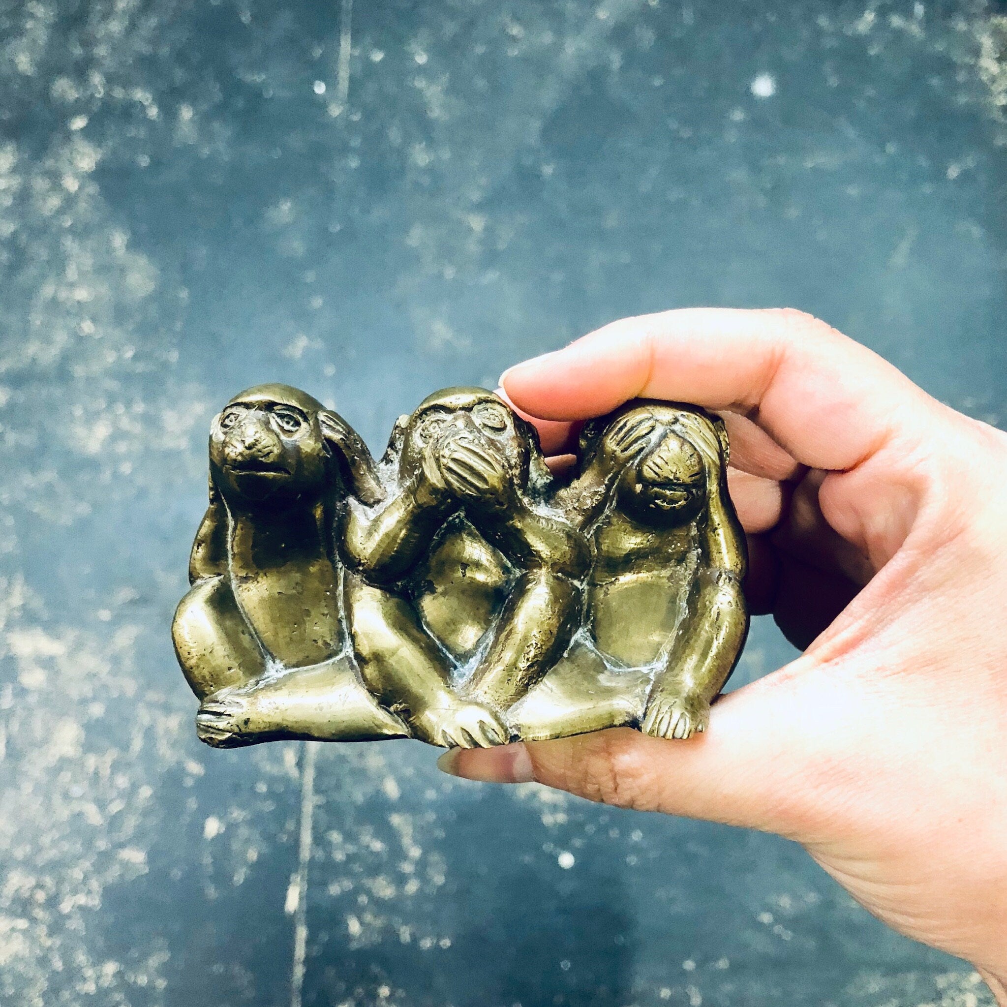 Hand holding brass figurine of three monkeys depicting "see no evil, hear no evil, speak no evil" proverb against blurred blue background