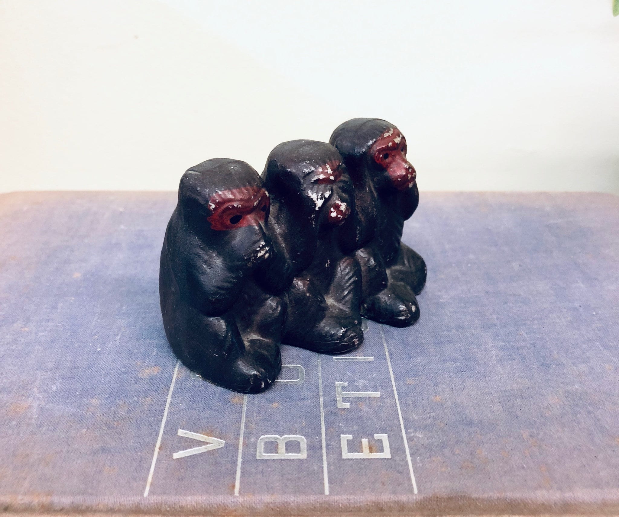 Three wise monkey figurines in black with red eyes, sitting in a row on a gray surface with measurement markings, depicting the "see no evil, hear no evil, speak no evil" proverb, vintage Japanese home decor trinket.