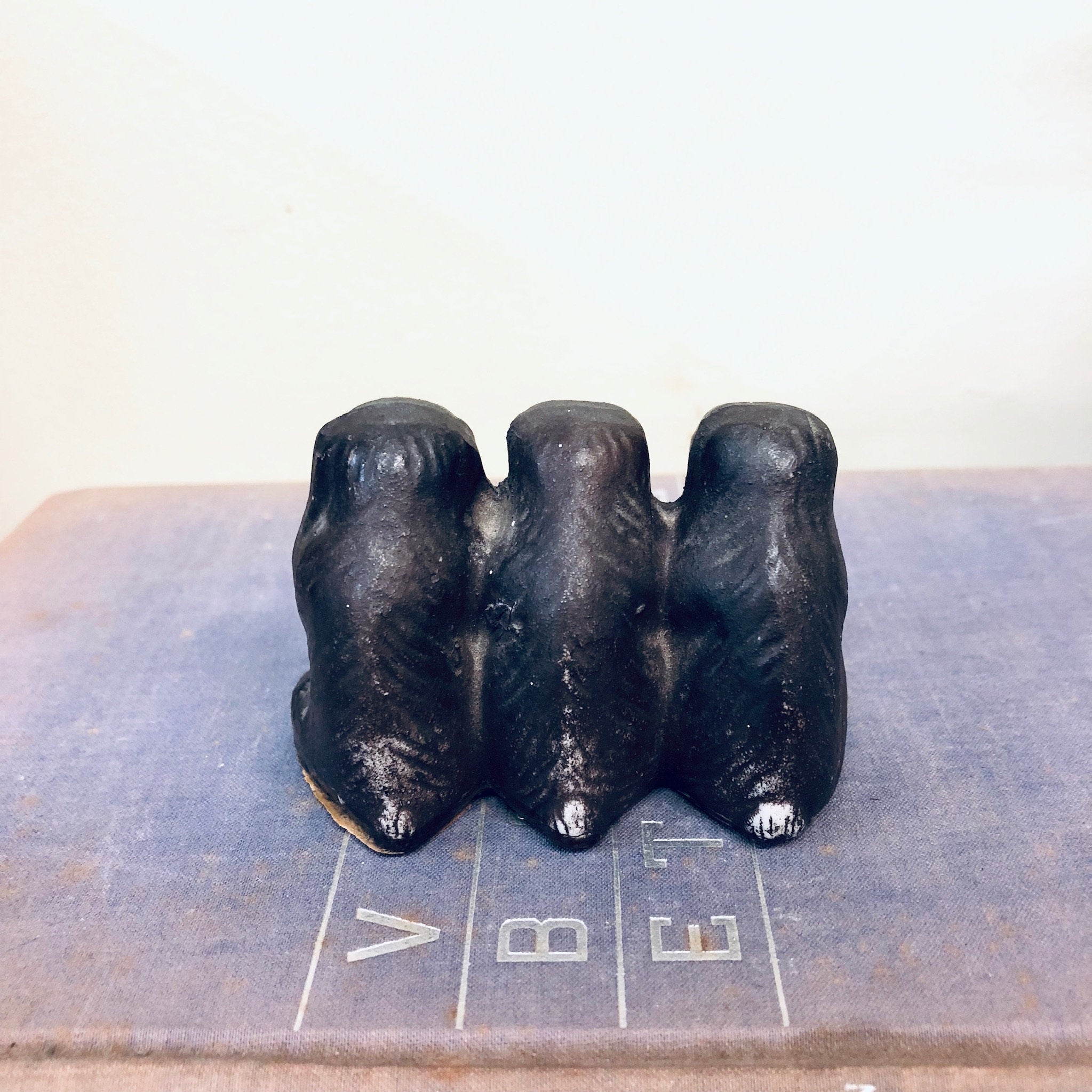 Three black monkey figurines sitting side by side on a gray surface, representing the "see no evil, hear no evil, speak no evil" proverb, with "V", "B", and "E" letters in front of each monkey