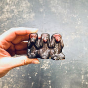Three ceramic monkey figurines depicting "see no evil, hear no evil, speak no evil" on a reflective surface being held in a hand.