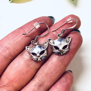 Vintage silver cat mask dangle earrings featuring cut-out design and cute kitty face details, shown in close-up view being held in a person's hand.