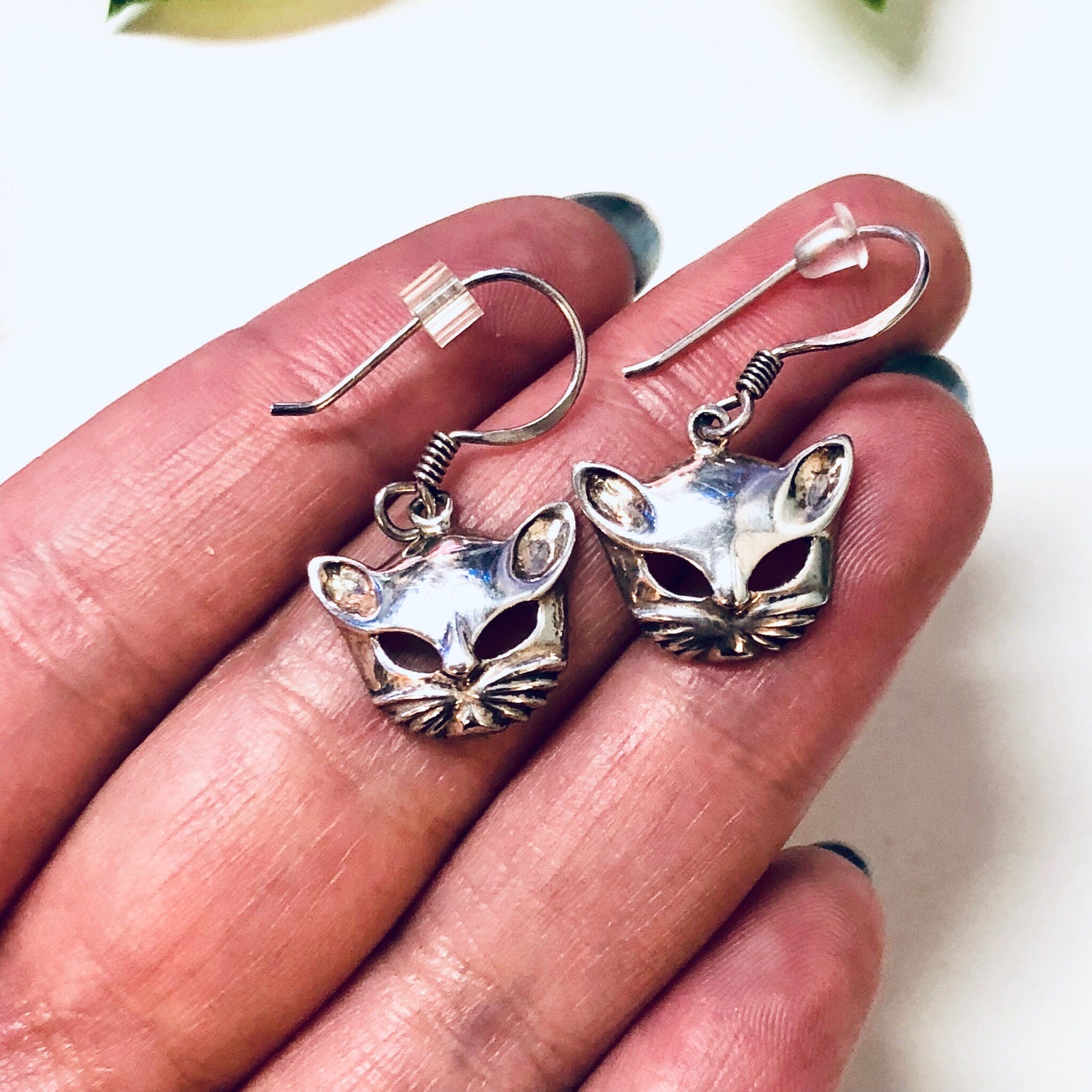 Vintage silver cat mask dangle earrings featuring cut-out design and cute kitty face details, shown in close-up view being held in a person's hand.