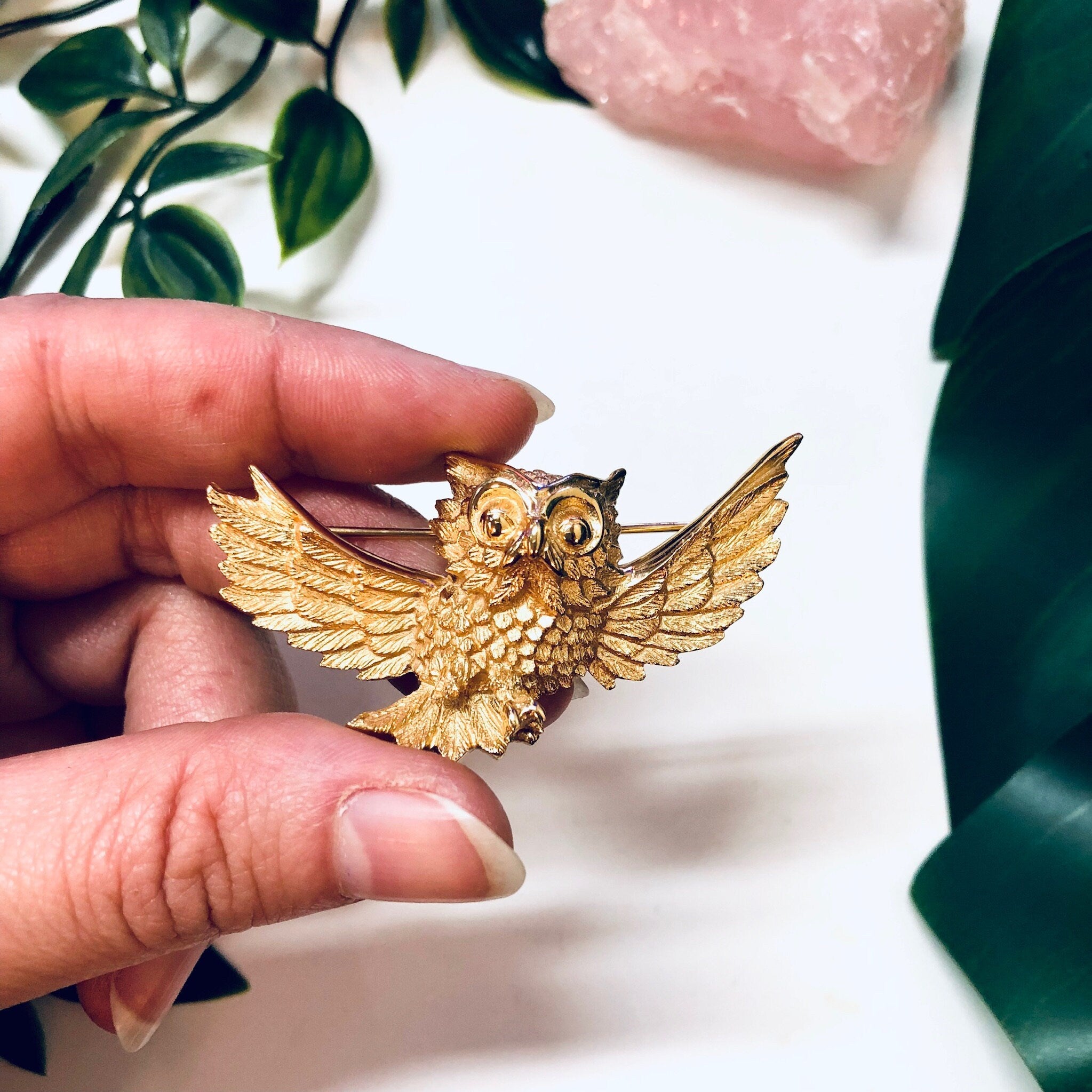 Vintage gold-toned Trifari owl brooch held in hand, with pink crystal and leaves in background.