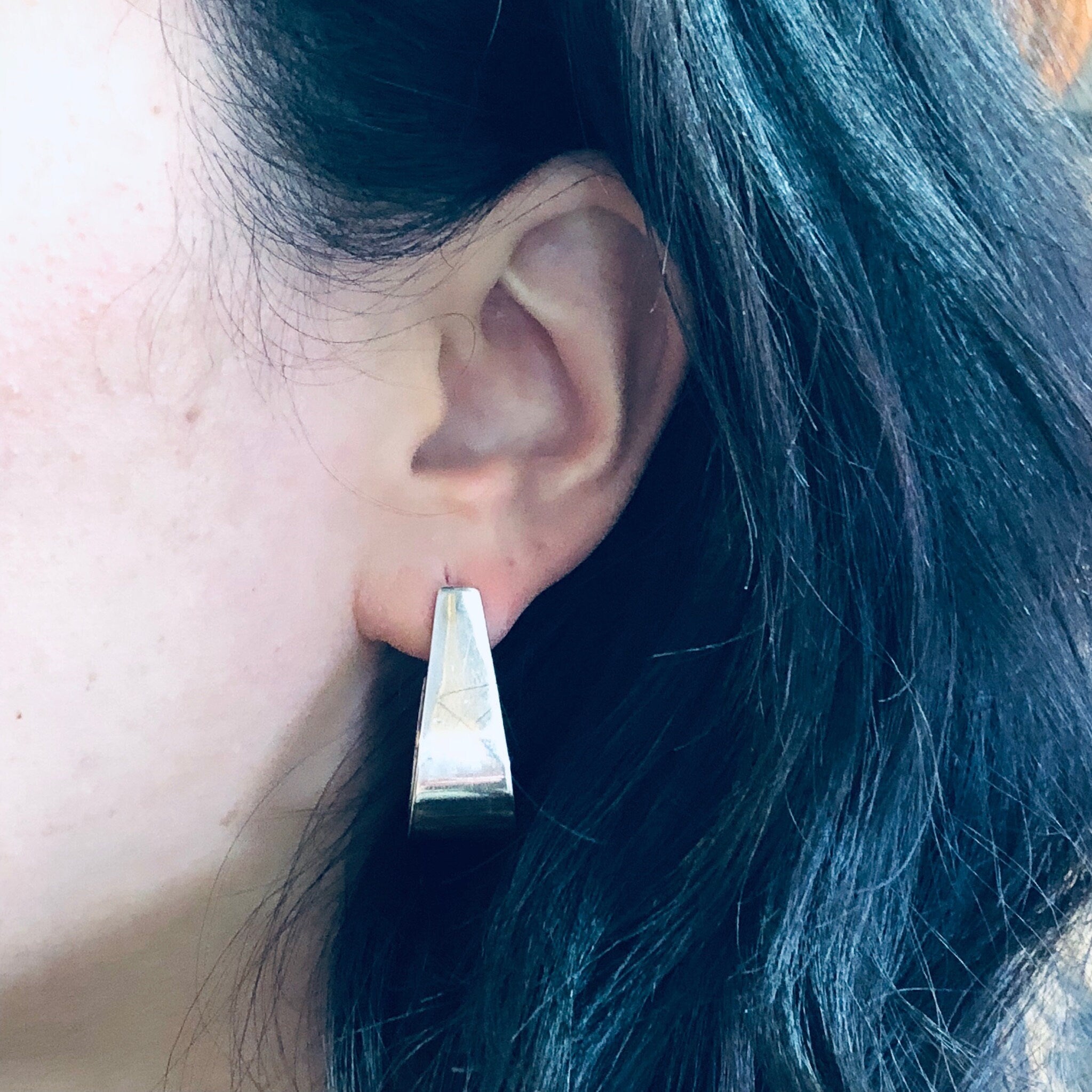 Close-up view of a person's ear wearing a shiny silver geometric earring against their dark hair. The earring has a thick, sculptural trapezoid shape.