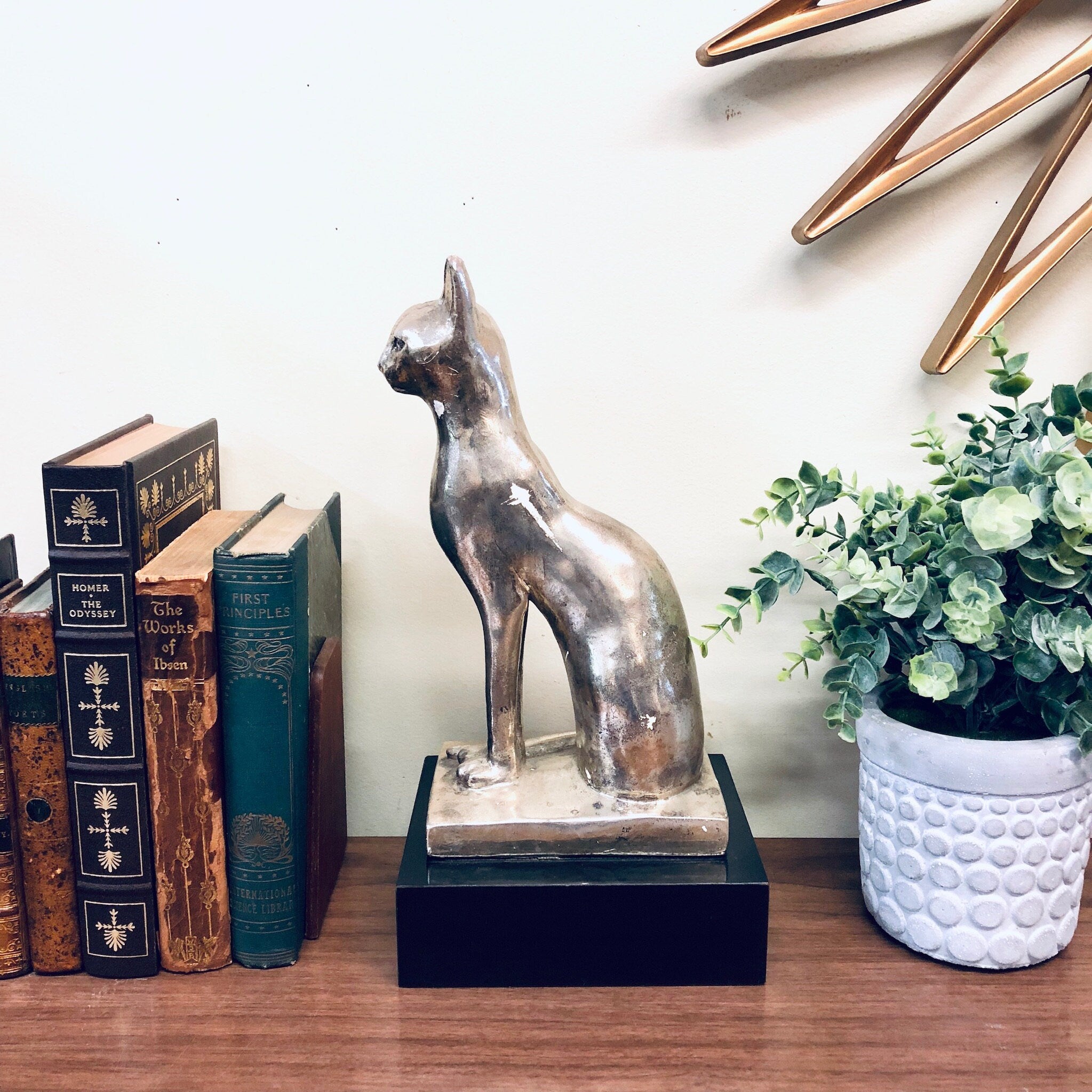 Silver cat statue on black stand next to vintage books and potted plant, modern home decor featuring animal sculpture
