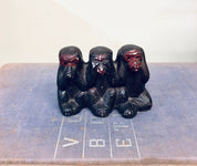 Three black ceramic monkey figurines covering their eyes, ears, and mouth, representing the "see no evil, hear no evil, speak no evil" proverb, sitting on a grid marked background.