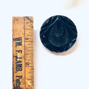 Round black carved Bakelite brooch from the 1930s next to a wooden ruler for scale, antique mourning jewelry pin.