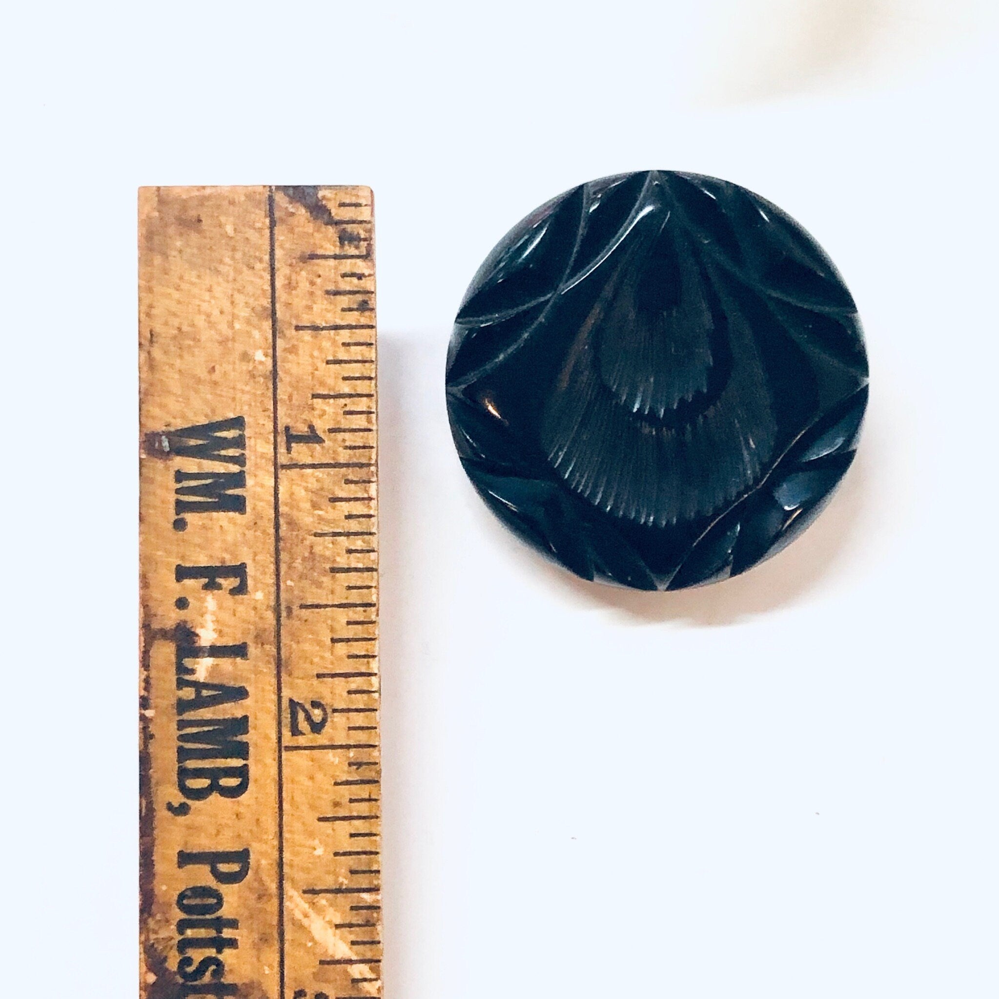 Round black carved Bakelite brooch from the 1930s next to a wooden ruler for scale, antique mourning jewelry pin.
