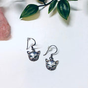 Vintage silver cat earrings with cut out kitty cat design, small dangle earrings resembling cat masks, cute cat jewelry displayed with leaves and crystals.