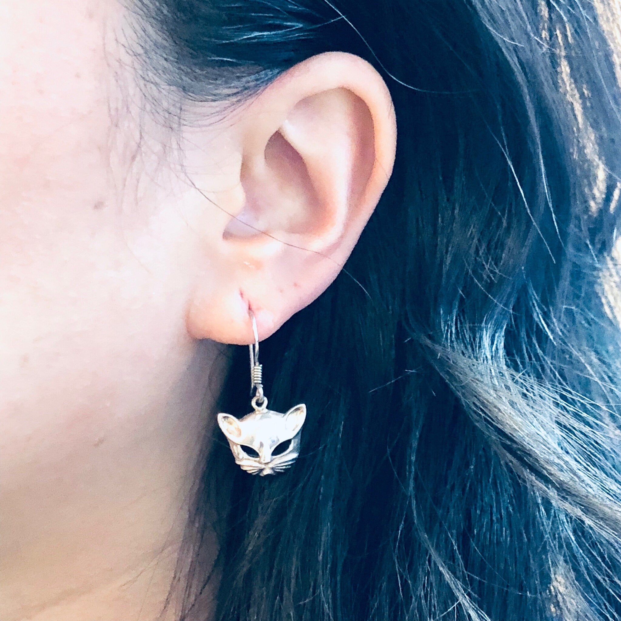 Close-up of a woman's ear wearing small silver cat head shaped dangle earrings against her dark hair.