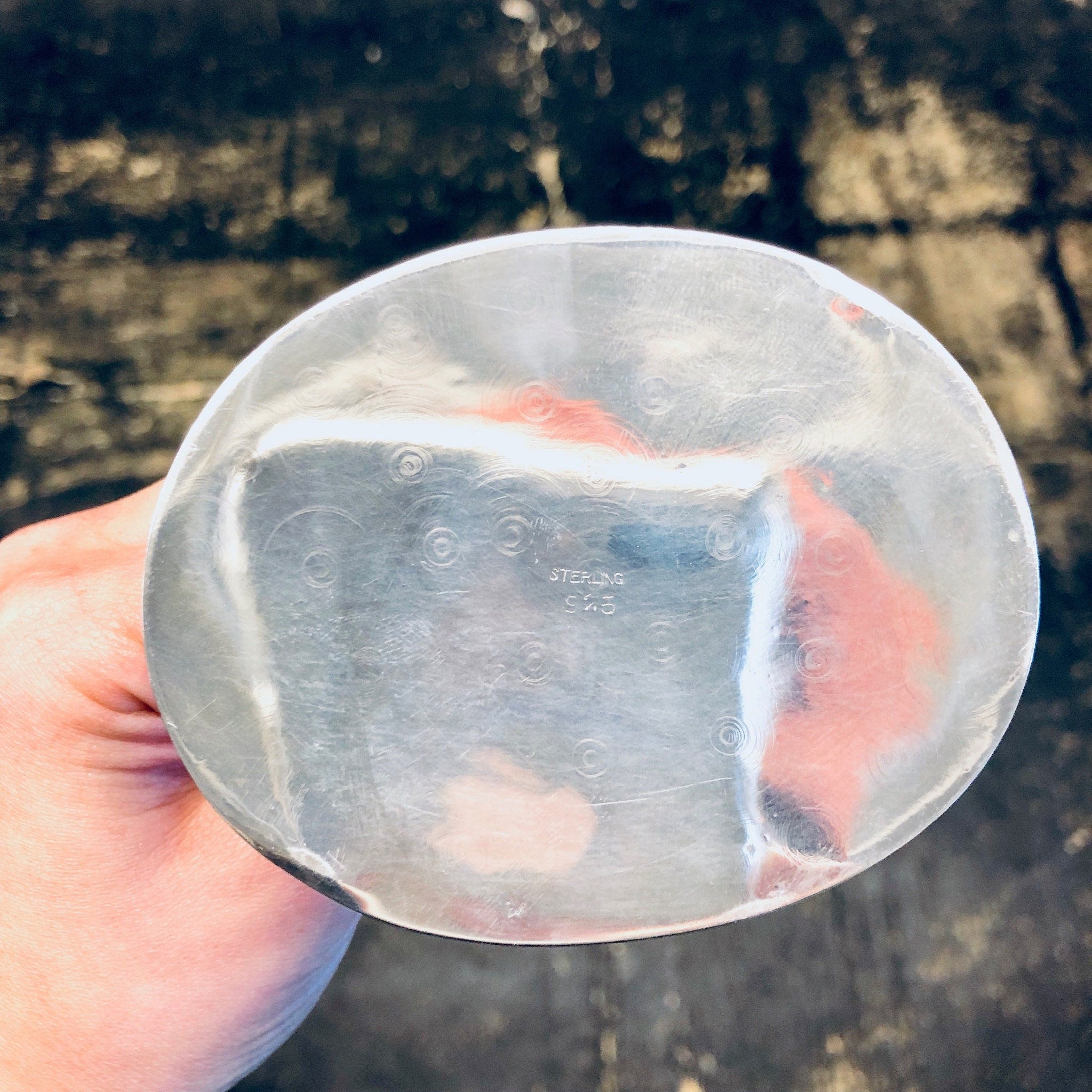 Hand holding an antique or vintage silver tray or plate that reflects sunlight, with a weathered stone or concrete background in soft focus.