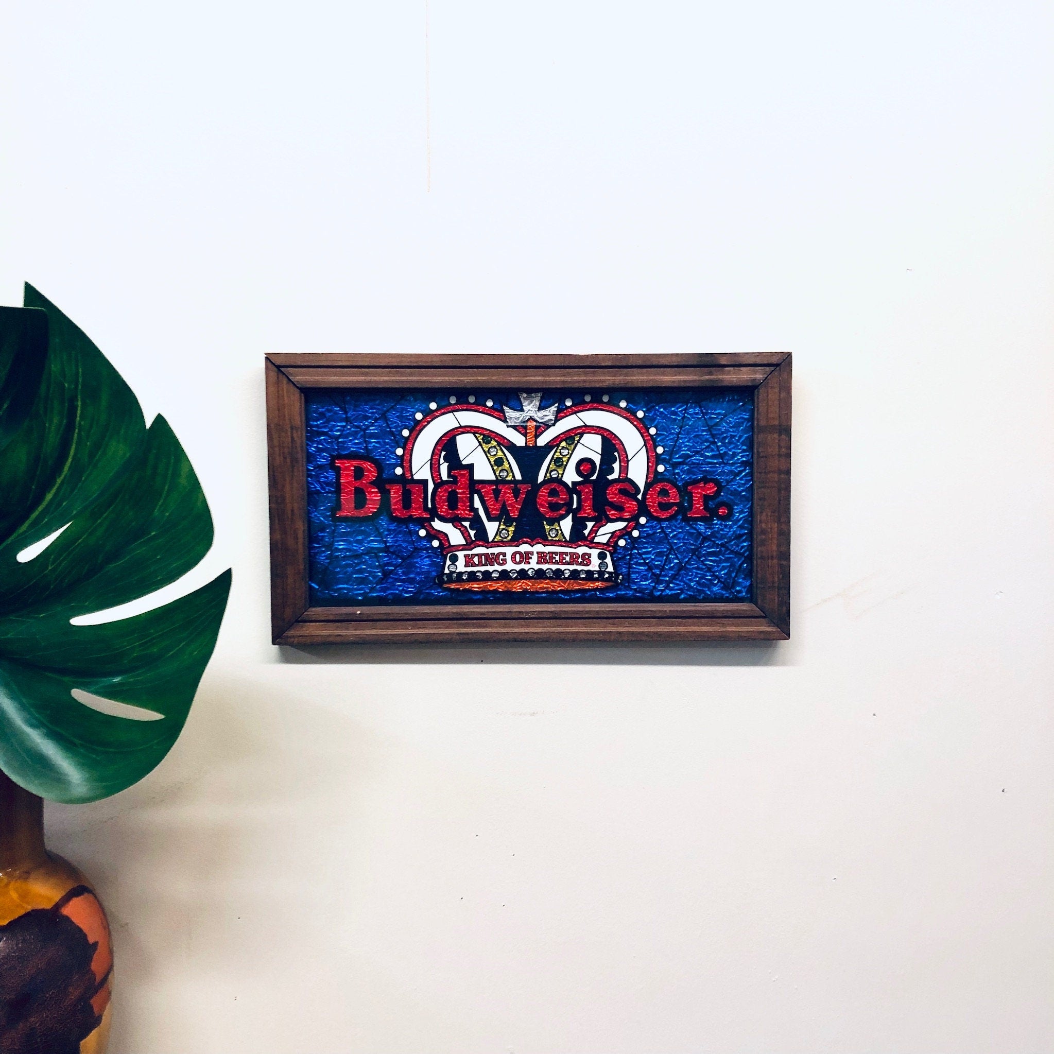 Vintage Budweiser "King of Beers" sign with crown logo in blue and red colors, framed in a wooden frame, hanging on a white wall next to a green plant leaf.