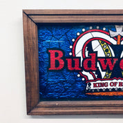 Vintage Budweiser beer sign featuring colorful blue background with red text and logo, framed in a rustic wooden frame, proclaiming Budweiser as the "King of Beers".
