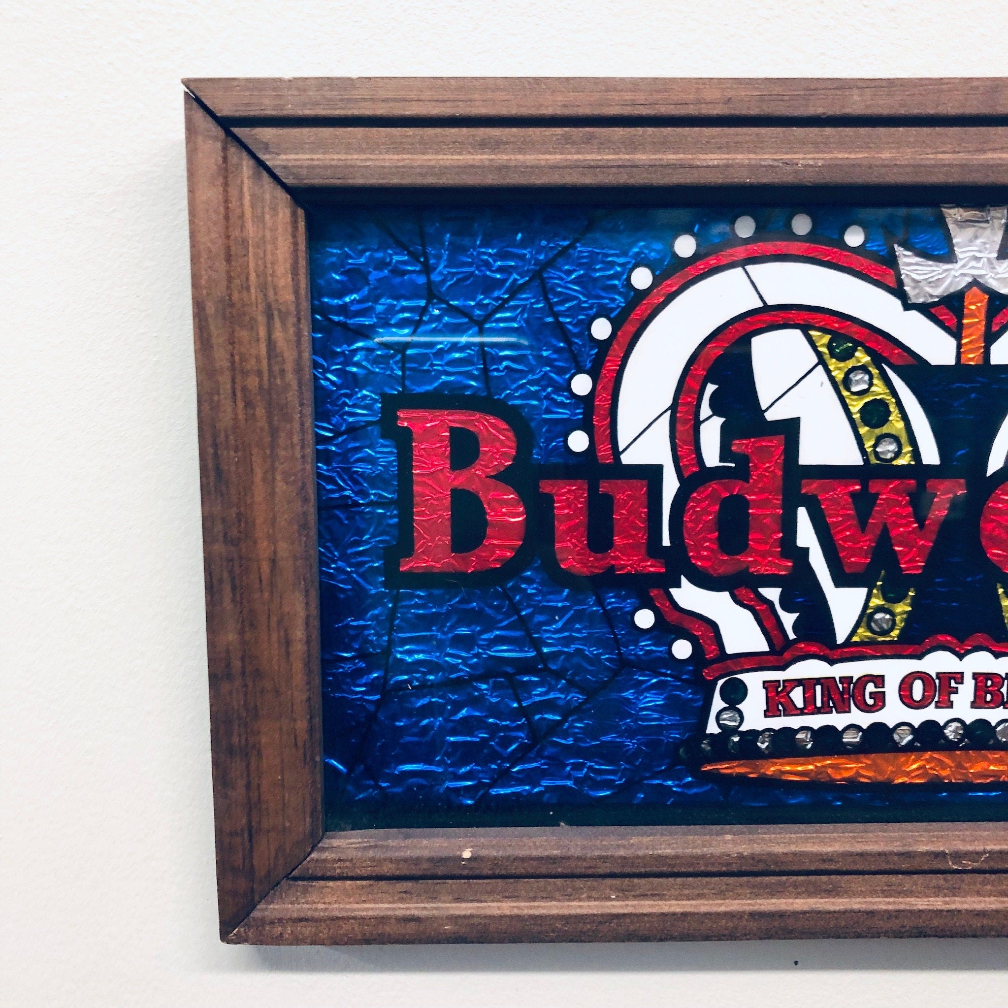 Vintage Budweiser beer sign featuring colorful blue background with red text and logo, framed in a rustic wooden frame, proclaiming Budweiser as the "King of Beers".