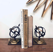 Vintage brass spherical sundial bookends with Roman numerals on a wooden shelf, surrounded by antique books. Unique home decor for a bookshelf or library.