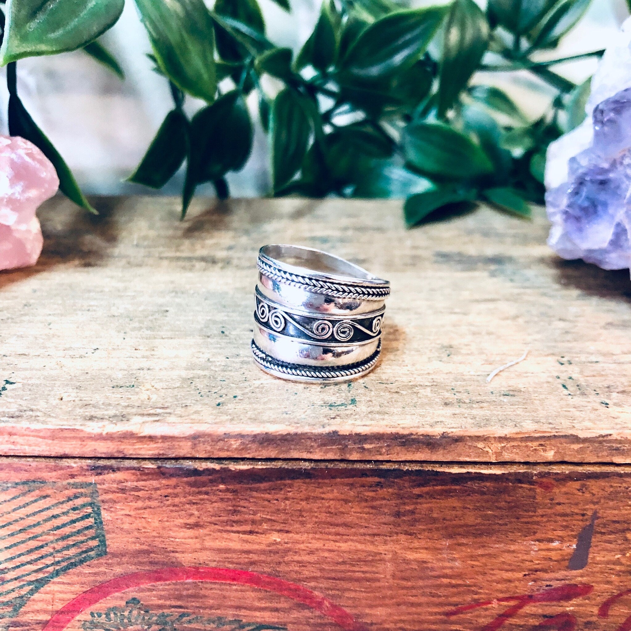 Vintage silver swirl pattern band ring on wooden surface with colorful crystals and greenery in background