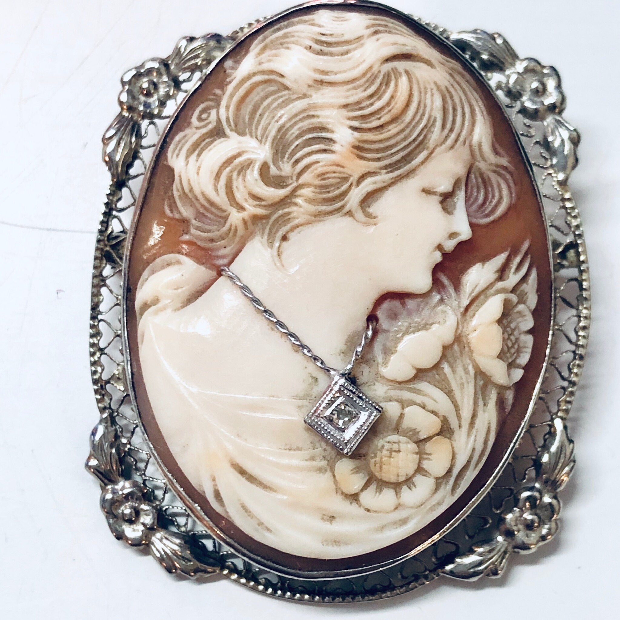 Vintage cameo brooch pendant featuring a carved shell portrait of a woman with flowing hair, set in an ornate 18K white gold frame adorned with floral and scroll motifs.