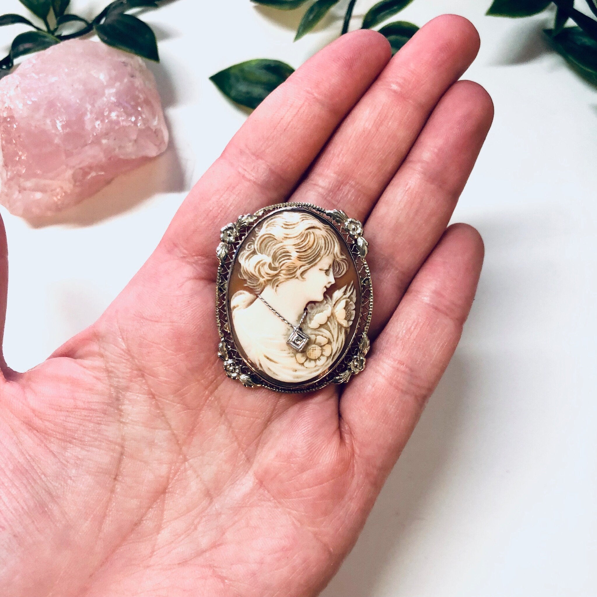 Vintage ornate cameo brooch pendant featuring a carved profile portrait set in an intricate 18K white gold frame, held in a person's hand against a white background with green foliage.