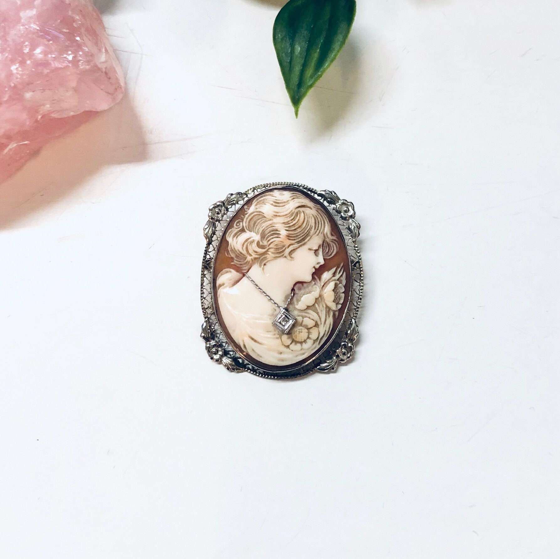 Ornate vintage cameo brooch pendant crafted in 18K white gold, featuring an intricately carved profile against a light background, surrounded by an elaborate floral frame with milgrain detailing, displayed on a white surface with pink crystal and green leaf accents.