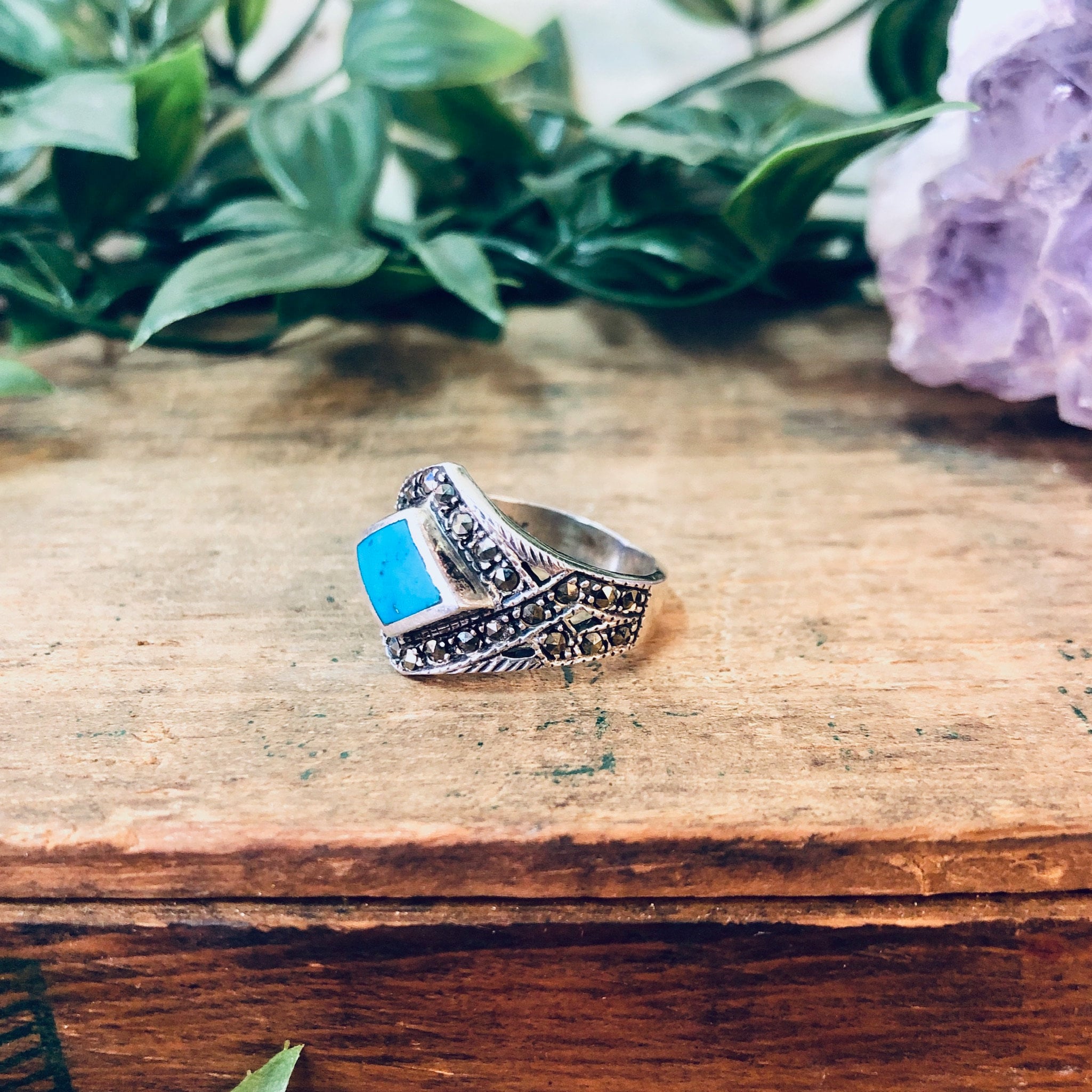 Vintage silver marcasite ring with turquoise stone set in a diamond shape, featuring sparkly details on a wooden surface with greenery in the background, stamped with 925 hallmark.