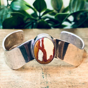 Vintage silver cuff bracelet with red natural stone inlay, set against a wooden surface with greenery in the background.