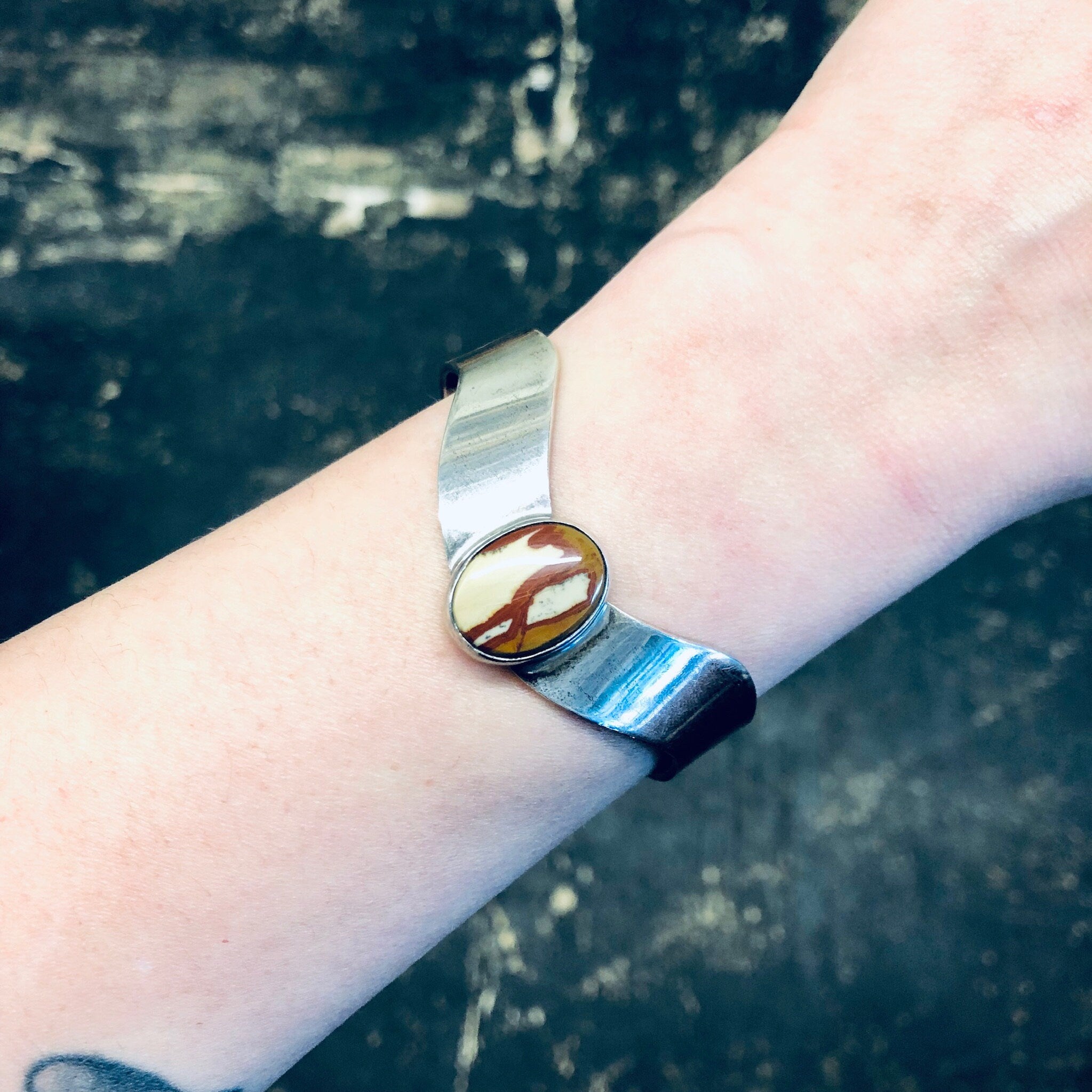 The image shows a vintage silver cuff bracelet with a red natural stone inlay worn on a person's wrist against an aged, weathered background.