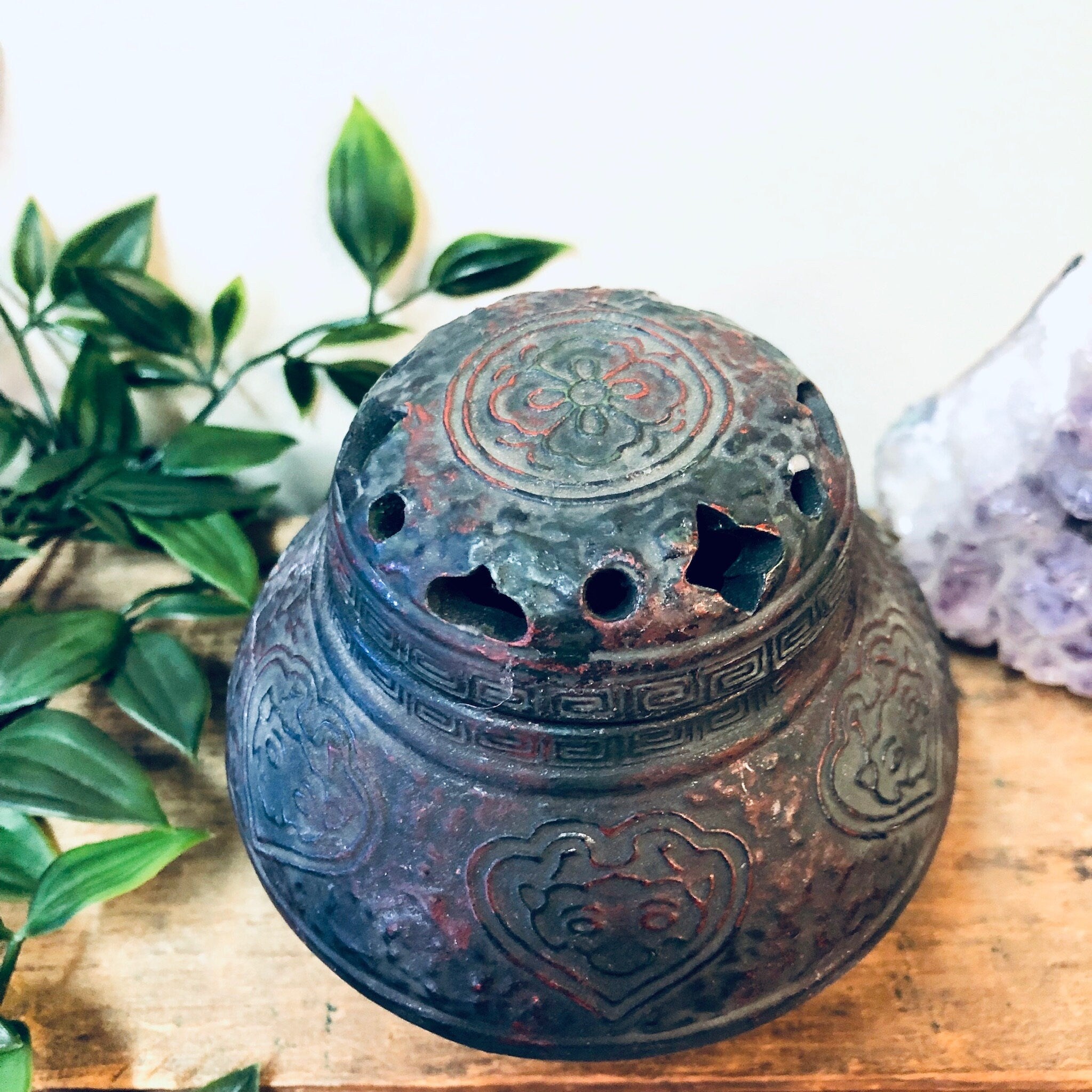 Antique bronze incense burner with intricate floral carvings, displayed with green foliage on a wooden surface, perfect as a unique housewarming gift or vintage home decor piece.