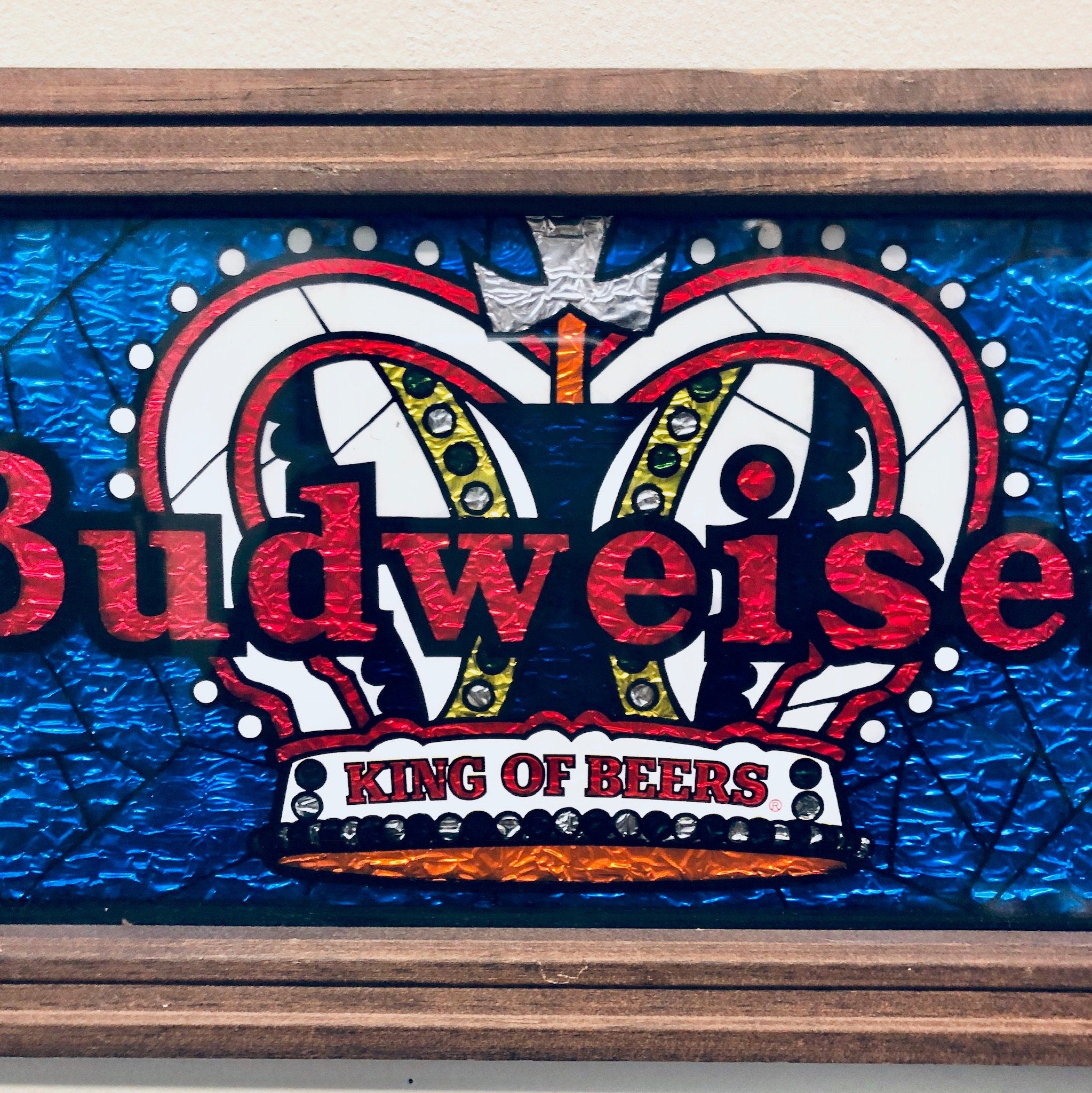 Vintage Budweiser beer sign with red and blue stained glass design featuring two rams and a crown, framed in wood with "King of Beers" text below the graphic.