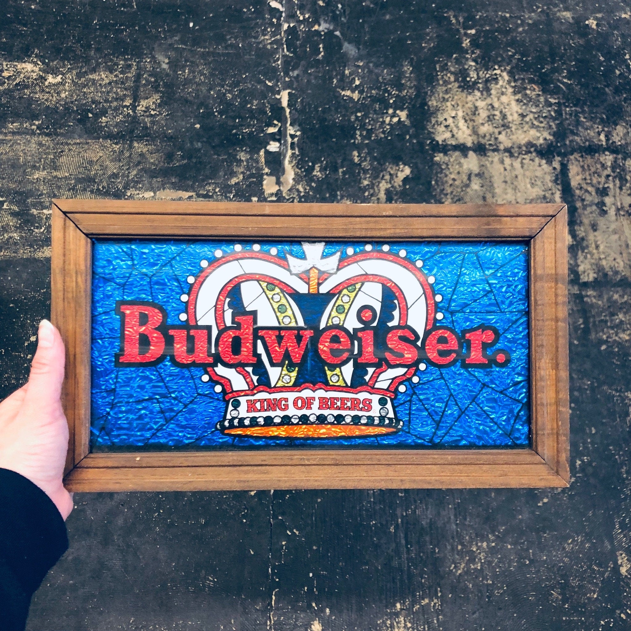 Vintage Budweiser "King of Beers" sign in blue and red, displayed in a wooden frame against a grungy wall.