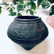 Antique embossed cast iron incense burner pot with floral heart design and green patina, surrounded by lush green foliage, perfect as a unique vintage home decor accent or housewarming gift.