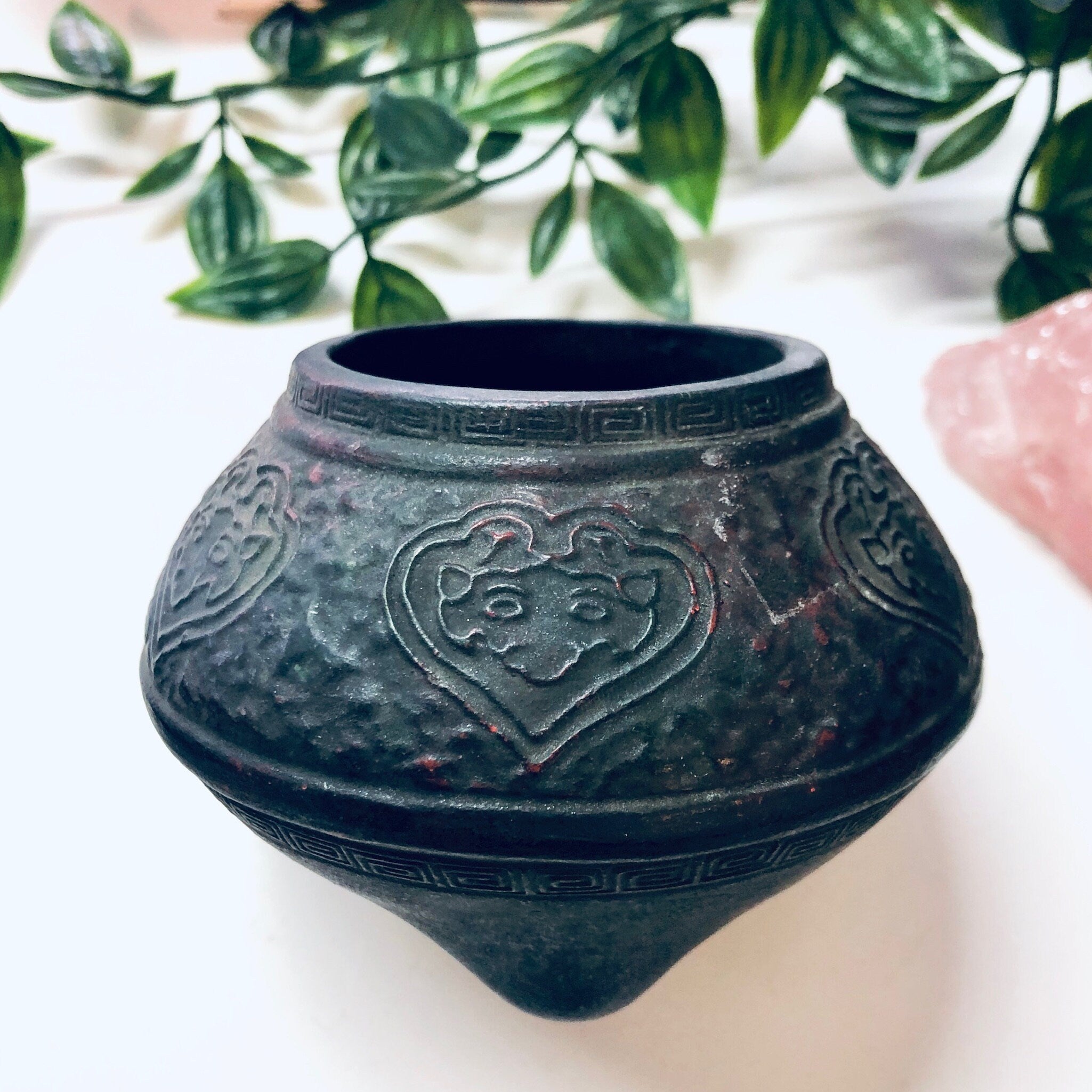 Antique embossed cast iron incense burner pot with floral heart design and green patina, surrounded by lush green foliage, perfect as a unique vintage home decor accent or housewarming gift.