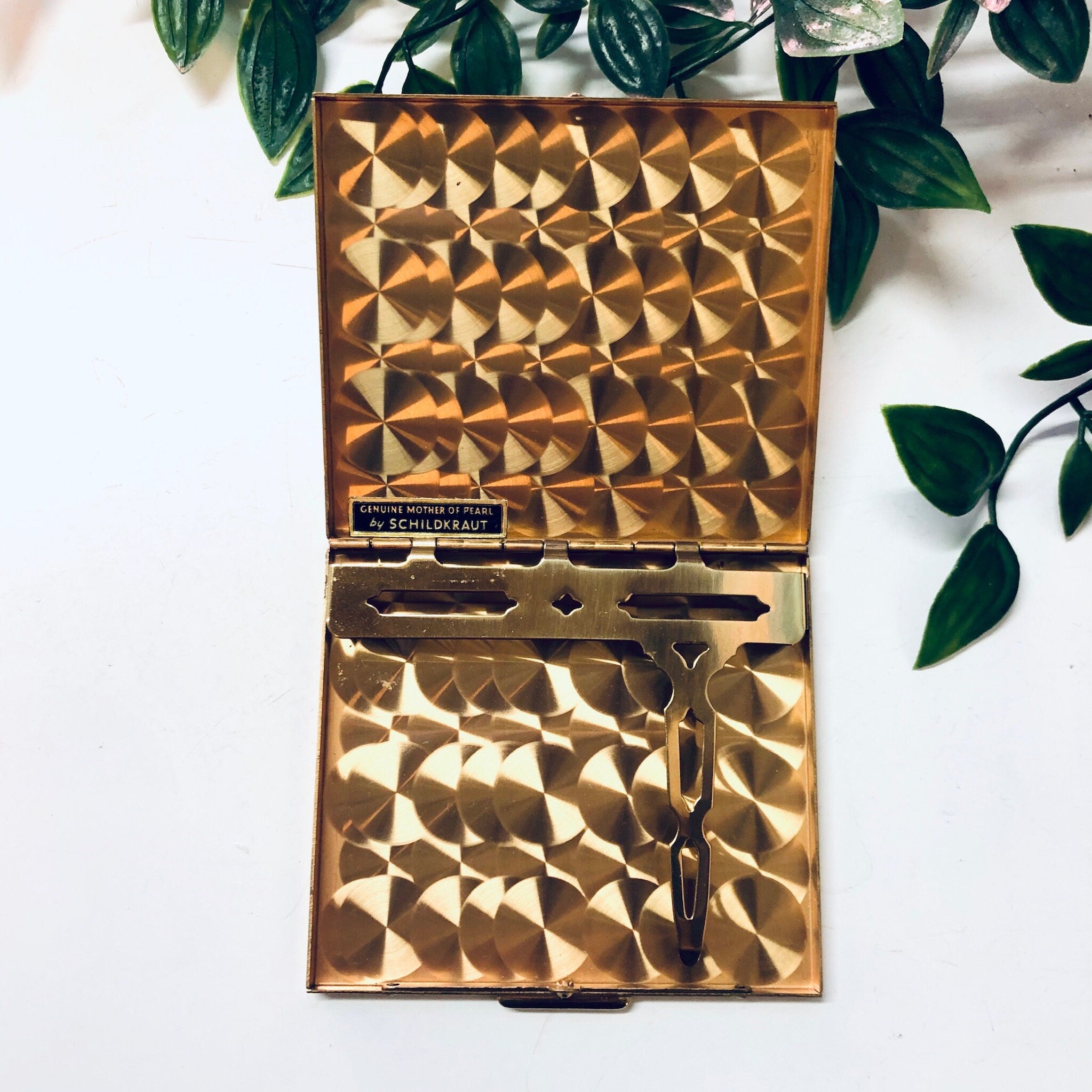 Vintage gold-tone metal cigarette case or business card holder with geometric mother of pearl inlay, made by Bailey Banks and Biddle, displayed open with razor blade on white fabric surrounded by green leaves.