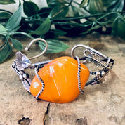 Vintage butterscotch amber and silver leaf cuff bracelet featuring a large orange amber stone and 925 silver details, displayed on a rustic wood surface with green foliage in the background.