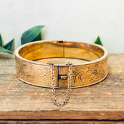 Vintage gold filled bangle bracelet with etched floral design and safety chain clasp, displayed on rustic wooden surface.