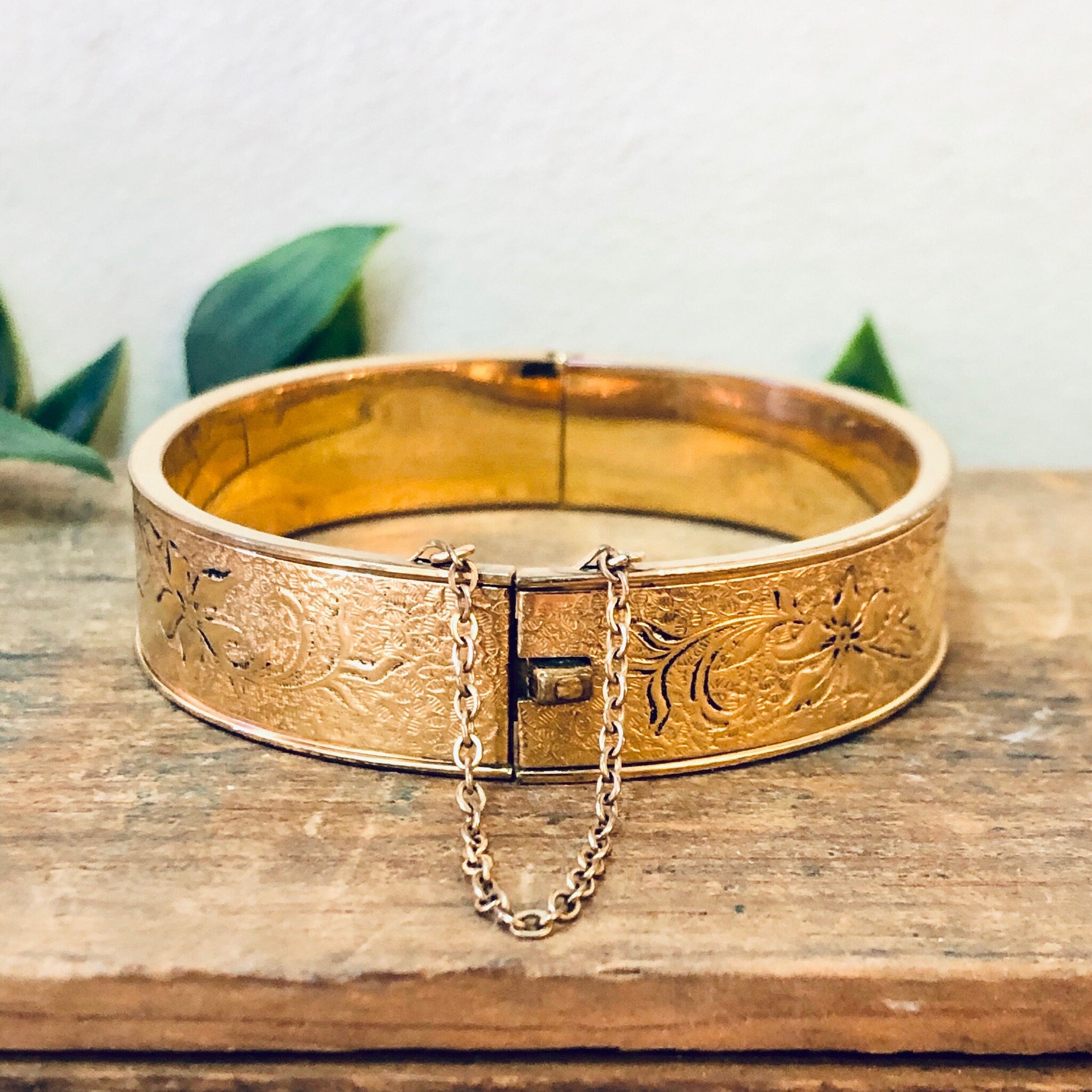 Vintage gold filled bangle bracelet with etched floral design and safety chain clasp, displayed on rustic wooden surface.